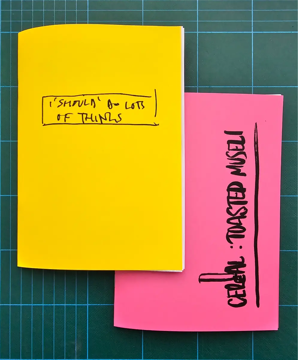 A yellow covered A5 zine and pink covered A5 zine sitting on a green cutting board.