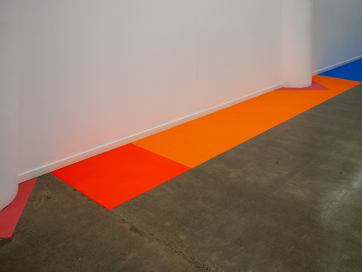 A short fluorescent orange rectangle, butts up to a long orange rectangle which in followed by a mid sized blue powder paint rectangle. All run along the edge of the wall. 